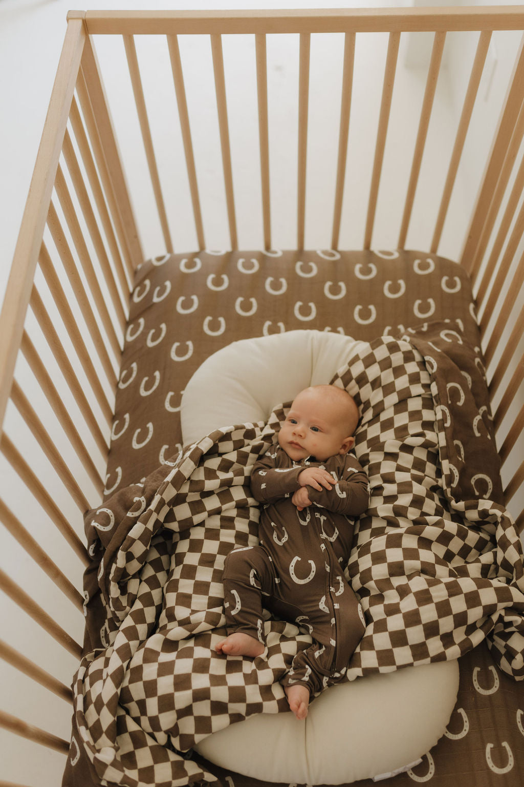 A baby relaxes in a wooden crib, snuggled under a cozy Muslin Quilt | Giddy Up/Dutton by forever french baby. The crib sheet features a brown and white checkerboard pattern, while the baby's outfit matches with delicate white horseshoes on brown fabric.