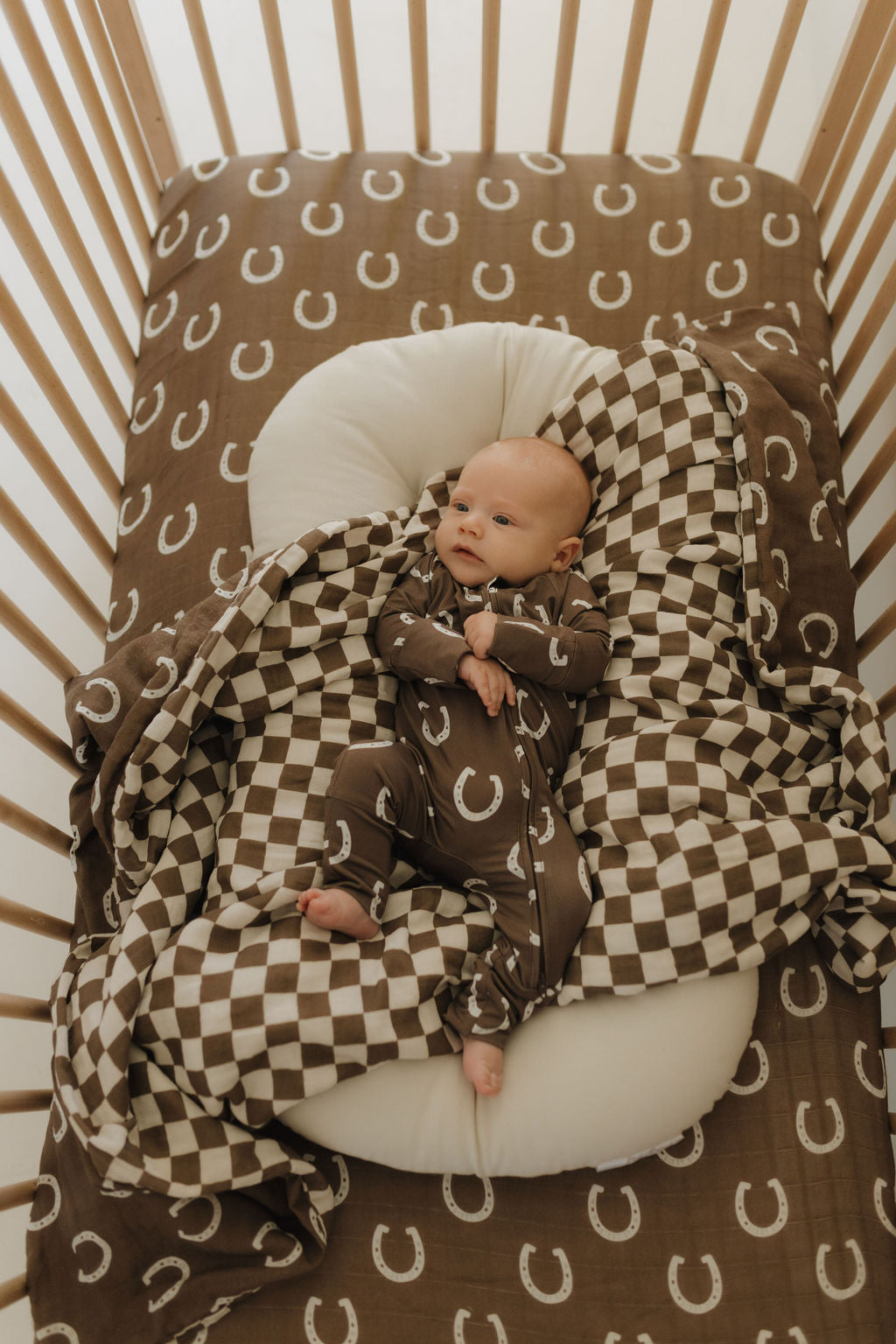 A baby in a crib lies on a cushion wrapped in a checkered blanket with horseshoe patterns. The crib's bedding matches for a cozy look. Wearing "Bamboo Zip Pajamas | Giddy Up" by forever french baby, the breathable fabric provides comfort and warmth with its brown horseshoe design.