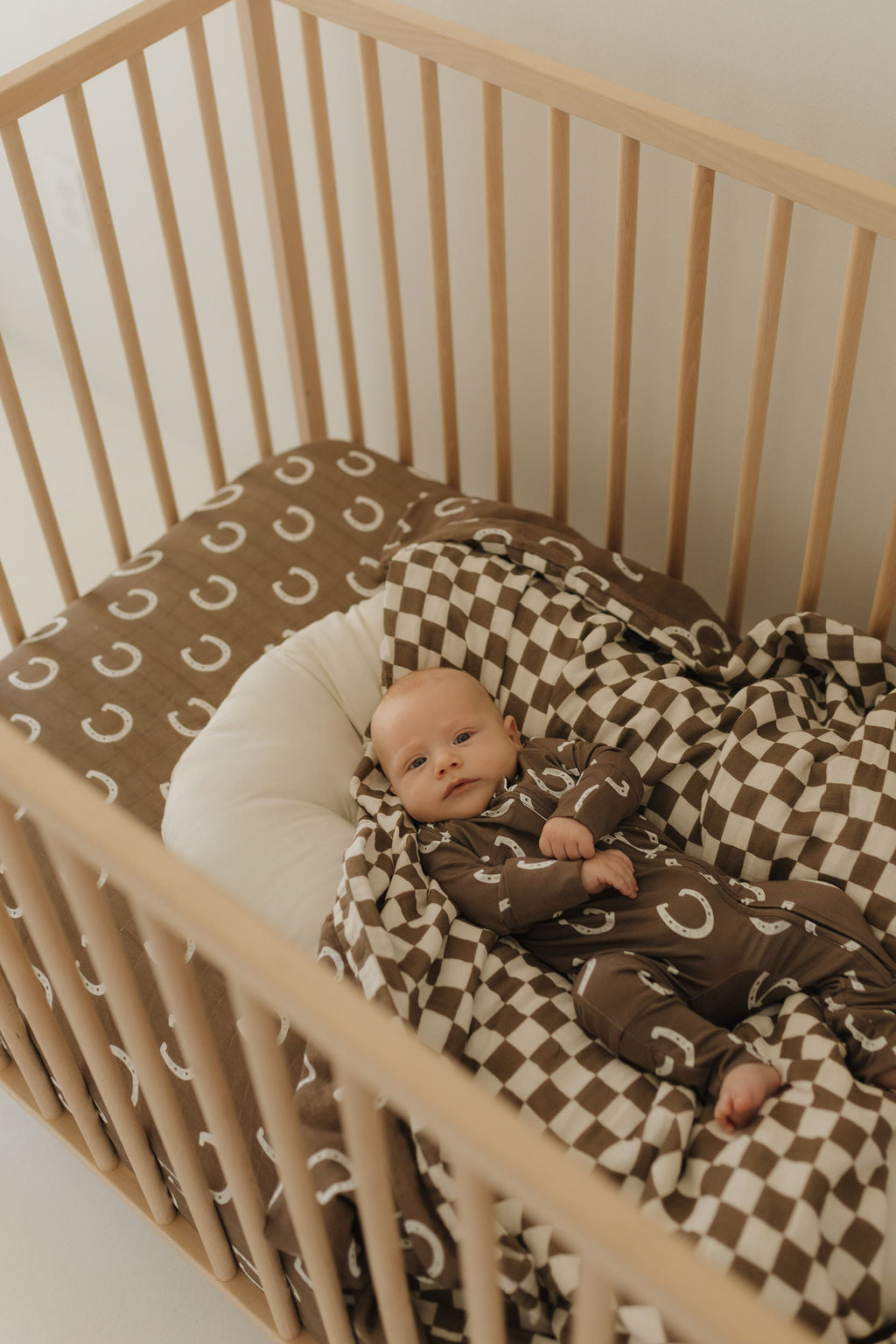 A baby rests in a wooden crib on a brown sheet with white crescents, wearing a matching outfit. Next to them is the Muslin Quilt | Giddy Up/Dutton by forever french baby, while a white cushioned pillow encircles their head, creating a cozy nursery atmosphere.