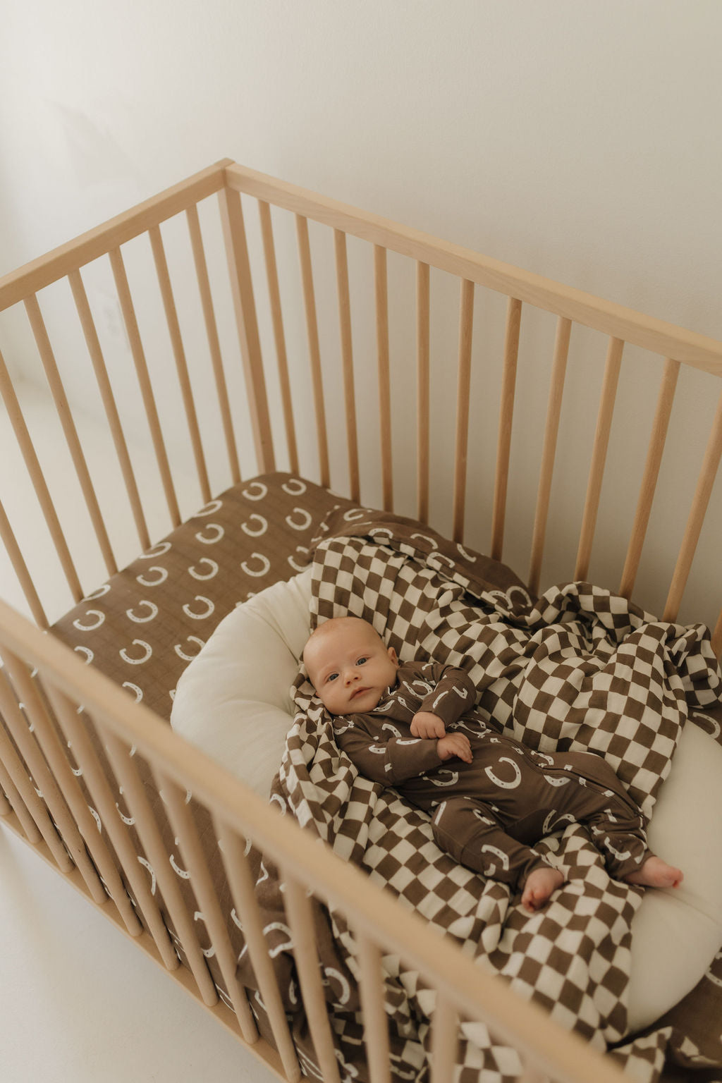 A baby in forever french baby's Bamboo Zip Pajamas | Giddy Up, featuring hypo-allergenic brown fabric with white crescents, lies relaxed in a wooden crib. The crib has a matching crescent-patterned brown blanket and a checkered one, as the baby gazes upwards.