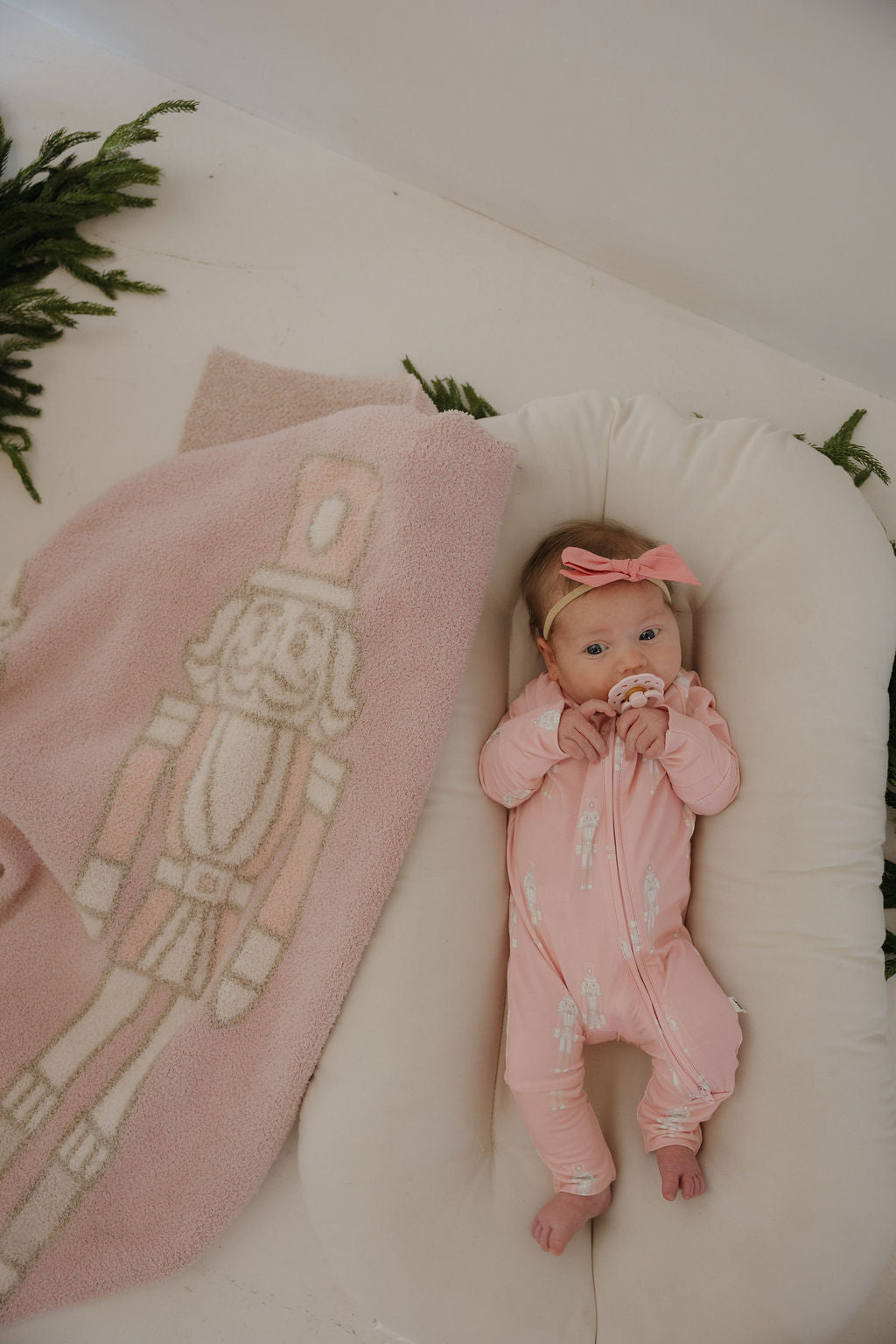 A baby in forever french baby's Bamboo Zip Pajamas | Nutcracker—a pink onesie—and headband lies in a cushioned lounger with a pacifier. A pink nutcracker blanket is nearby, greenery decorates the background.