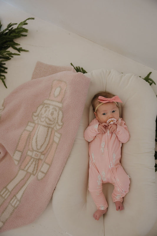 A baby in forever french baby's Bamboo Zip Pajamas | Nutcracker—a pink onesie—and headband lies in a cushioned lounger with a pacifier. A pink nutcracker blanket is nearby, greenery decorates the background.