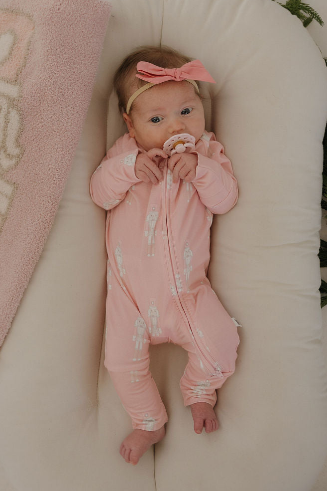 A baby in forever french baby's Bamboo Zip Pajamas | Nutcracker and a matching headband lounges on a cream cushion, cozy and hypoallergenic, gazing up with a pacifier in their mouth.