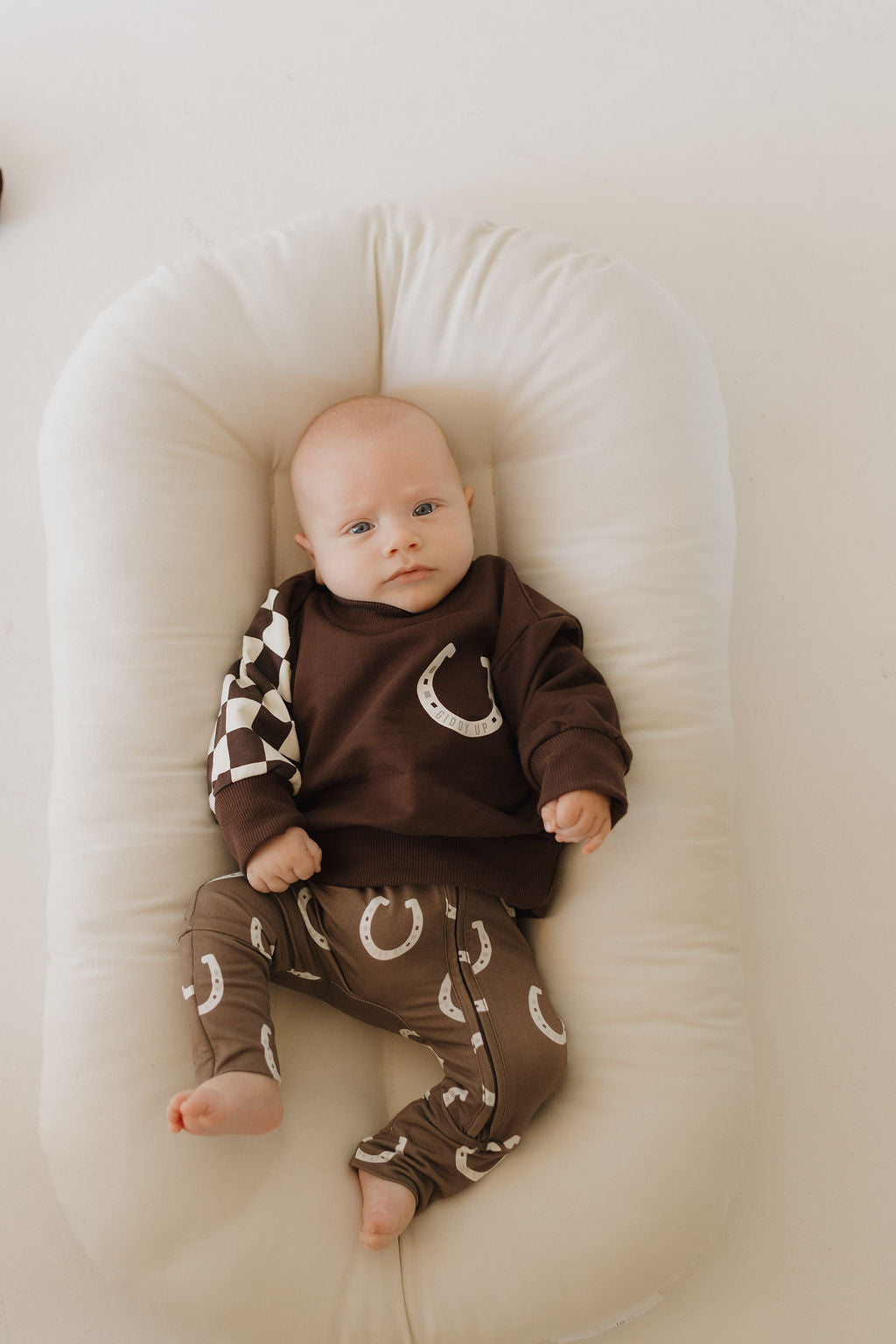 A baby lies on a soft, cream-colored cushion, wrapped in forever french baby's Bamboo Zip Pajamas | Giddy Up. The hypo-allergenic clothing features horseshoe and checkered patterns on breathable fabric, ensuring comfort against the plain, neutral background.