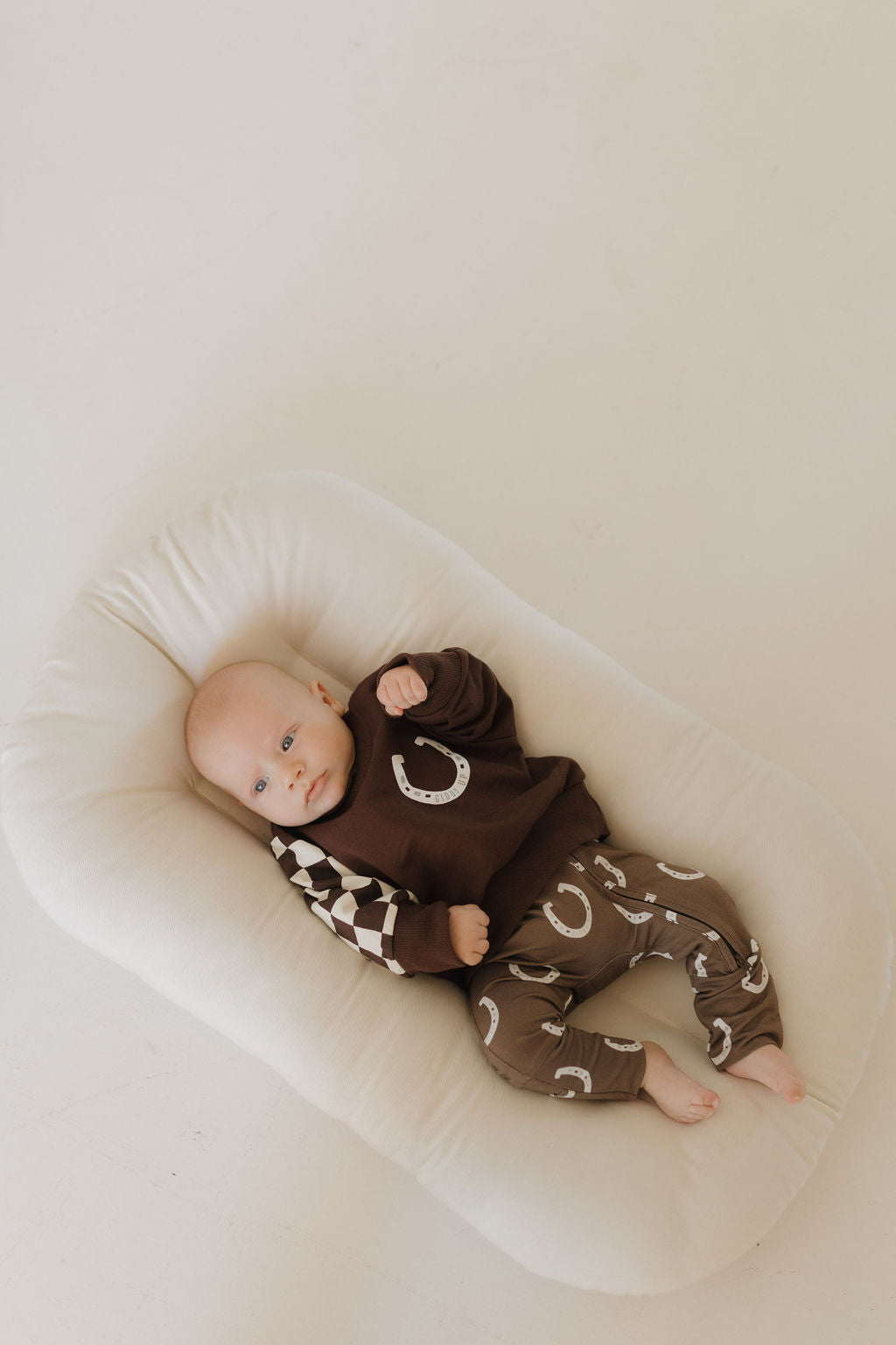 A calm-looking baby lies on a white cushion in hypo-allergenic Bamboo Zip Pajamas | Giddy Up by forever french baby, featuring a brown outfit with crescent designs and checkered sleeves, nestled against a plain light background.