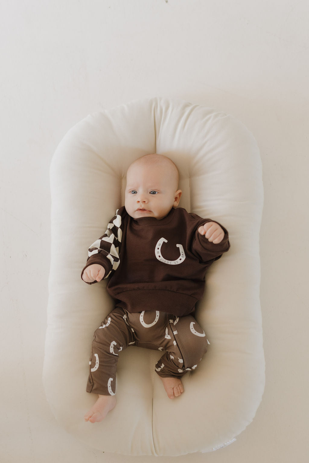 A baby lies on a soft white cushion, wearing breathable hypo-allergenic "Giddy Up" Bamboo Zip Pajamas by forever french baby. The calming brown outfit features horseshoe patterns and a black-and-white checkered sleeve as the baby's gaze is directed upward with a relaxed expression.