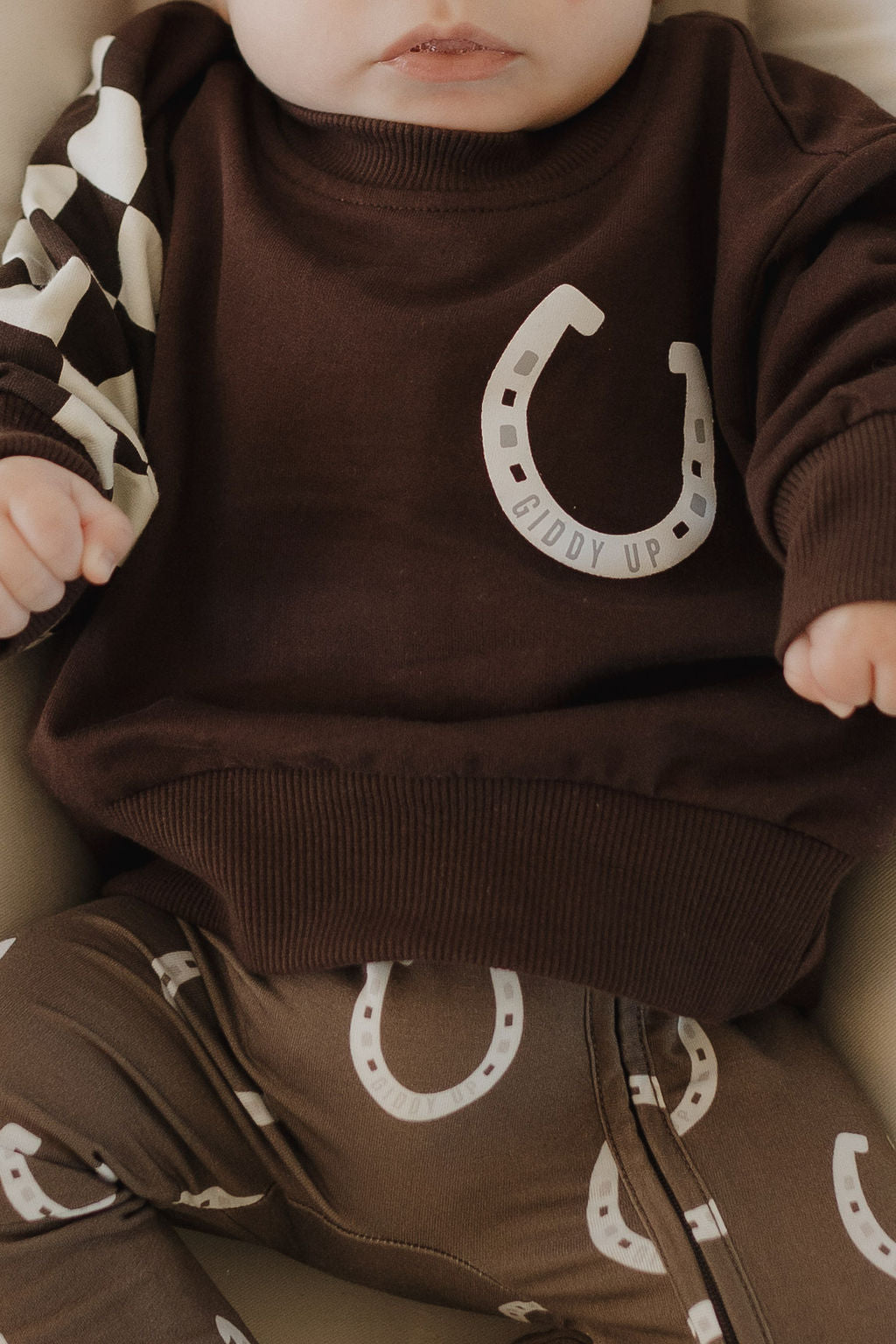 A baby wears forever french baby's Bamboo Zip Pajamas in brown with horseshoe prints and a "Giddy Up" design on the chest. Featuring a hypo-allergenic, breathable blend, the shirt includes a sleeve with a white and brown pattern, ensuring comfort while seated and visible from the torso up.