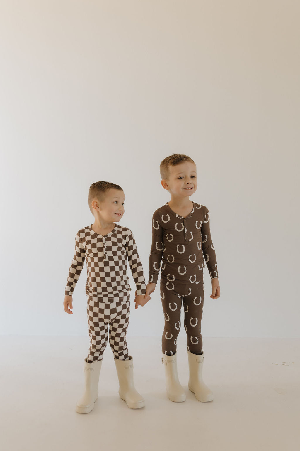 Two children hold hands, smiling sweetly. One wears matching white boots with a brown and white checkered outfit, the other in "Bamboo Two Piece Pajamas | Dutton" by forever french baby, featuring white horseshoe patterns. They exude comfort and style against a plain background.