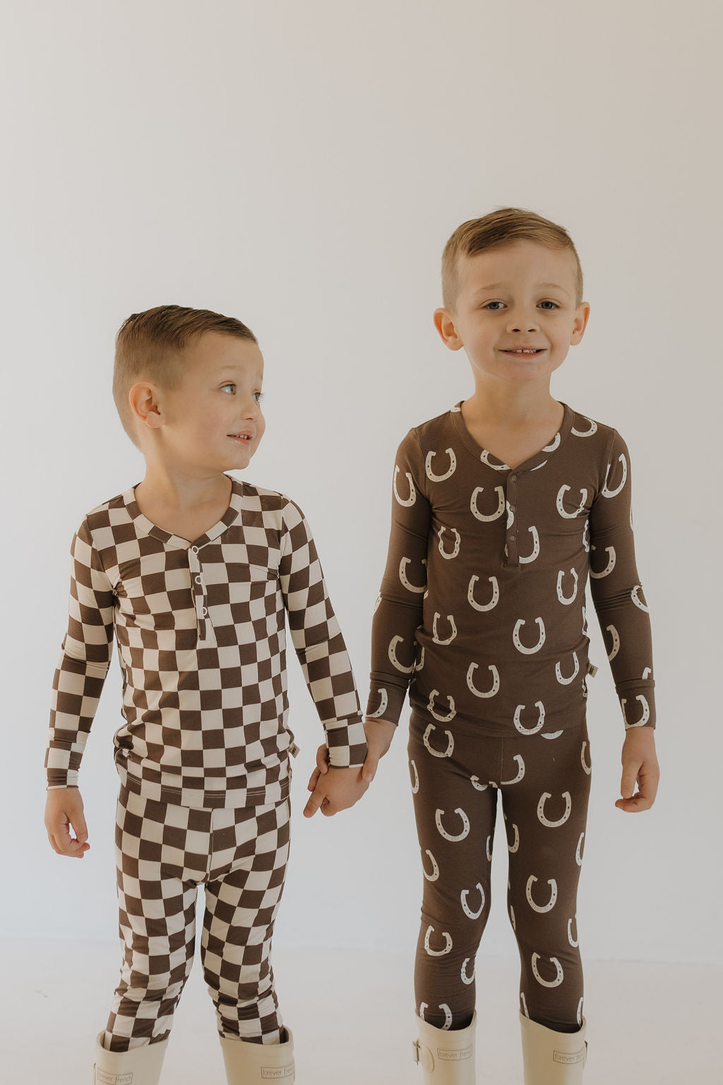 Two young boys stand hand in hand, smiling warmly in their breathable Bamboo Two Piece Pajamas from forever french baby. One sports a brown and white checkered pattern, the other wears pajamas adorned with white horseshoes, both pairing them with cozy beige boots.