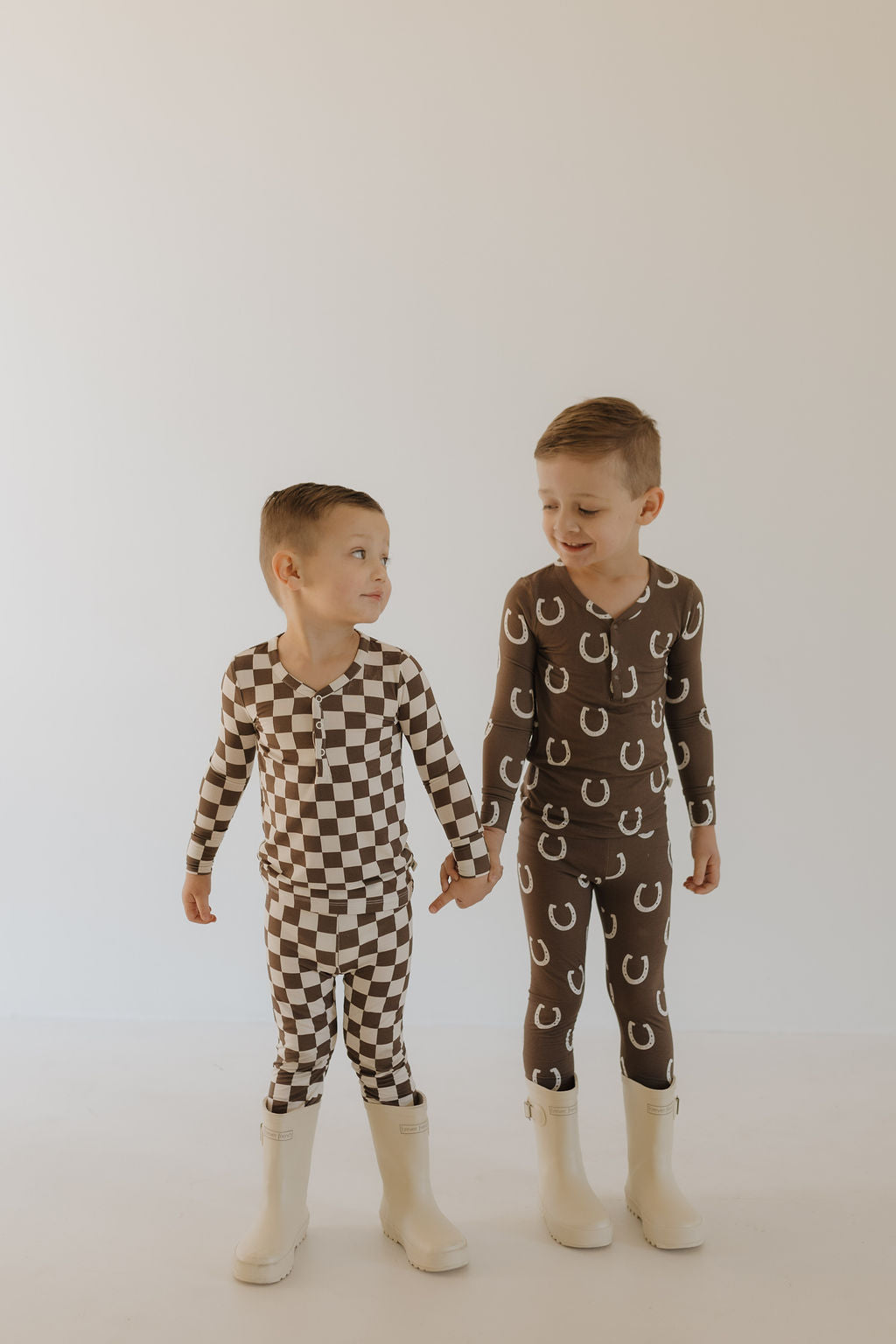 In a neutral-colored room, two young children holding hands beam with happiness in cozy outfits. One sports a brown and white checkered look, the other wears charming brown pajamas with white horseshoes from forever french baby, possibly hypo-allergenic Bamboo Two Piece Pajamas | Dutton.