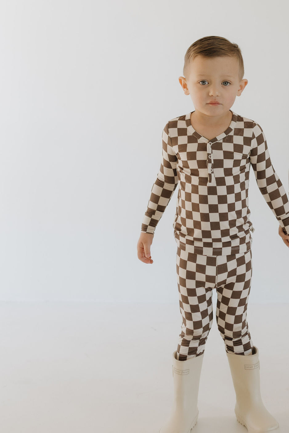 A young child stands against a plain background in the Bamboo Two Piece Pajamas | Dutton from forever french baby, featuring a brown and white checkered design with long sleeves and pants. Beige boots complete the look as the child showcases the comfort of breathable sleepwear.