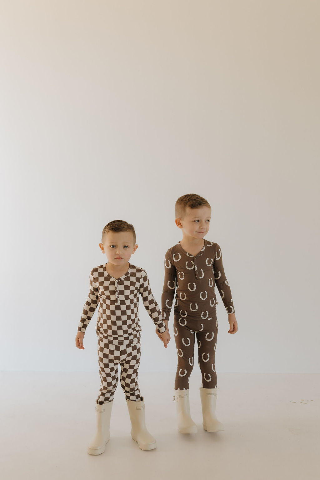 Two young children hold hands in Forever French Baby's breathable Bamboo Two Piece Pajamas | Dutton. One sports a brown checkered pattern, the other wears brown with white horseshoe prints. Both have short hair and wear white boots against a plain light background.