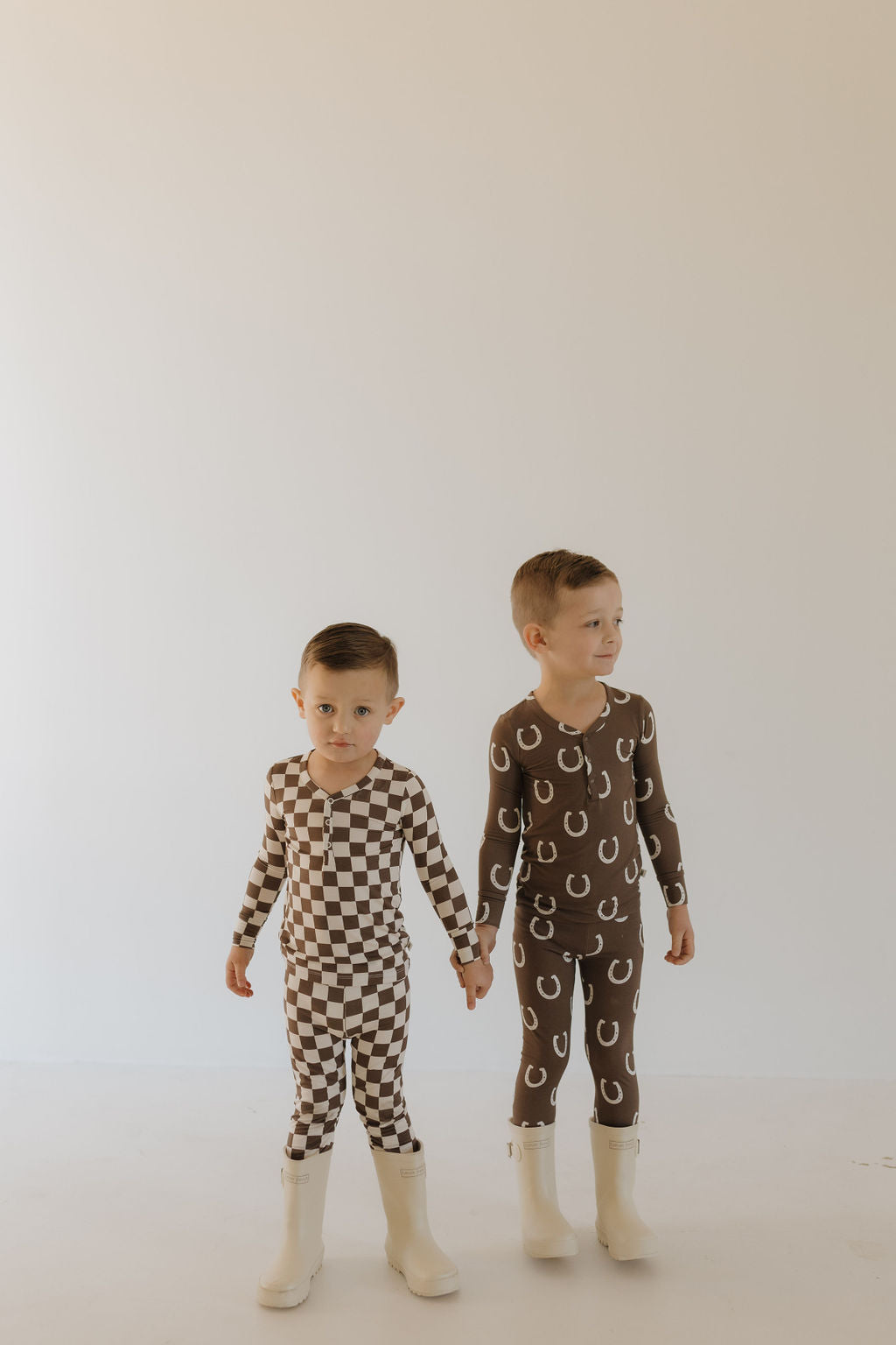 Two young children hold hands, wearing charming breathable sleepwear from forever french baby. One dons brown and white checkered pajamas while the other wears an outfit with playful horseshoe patterns. Both sport white boots against a plain, light-colored background.