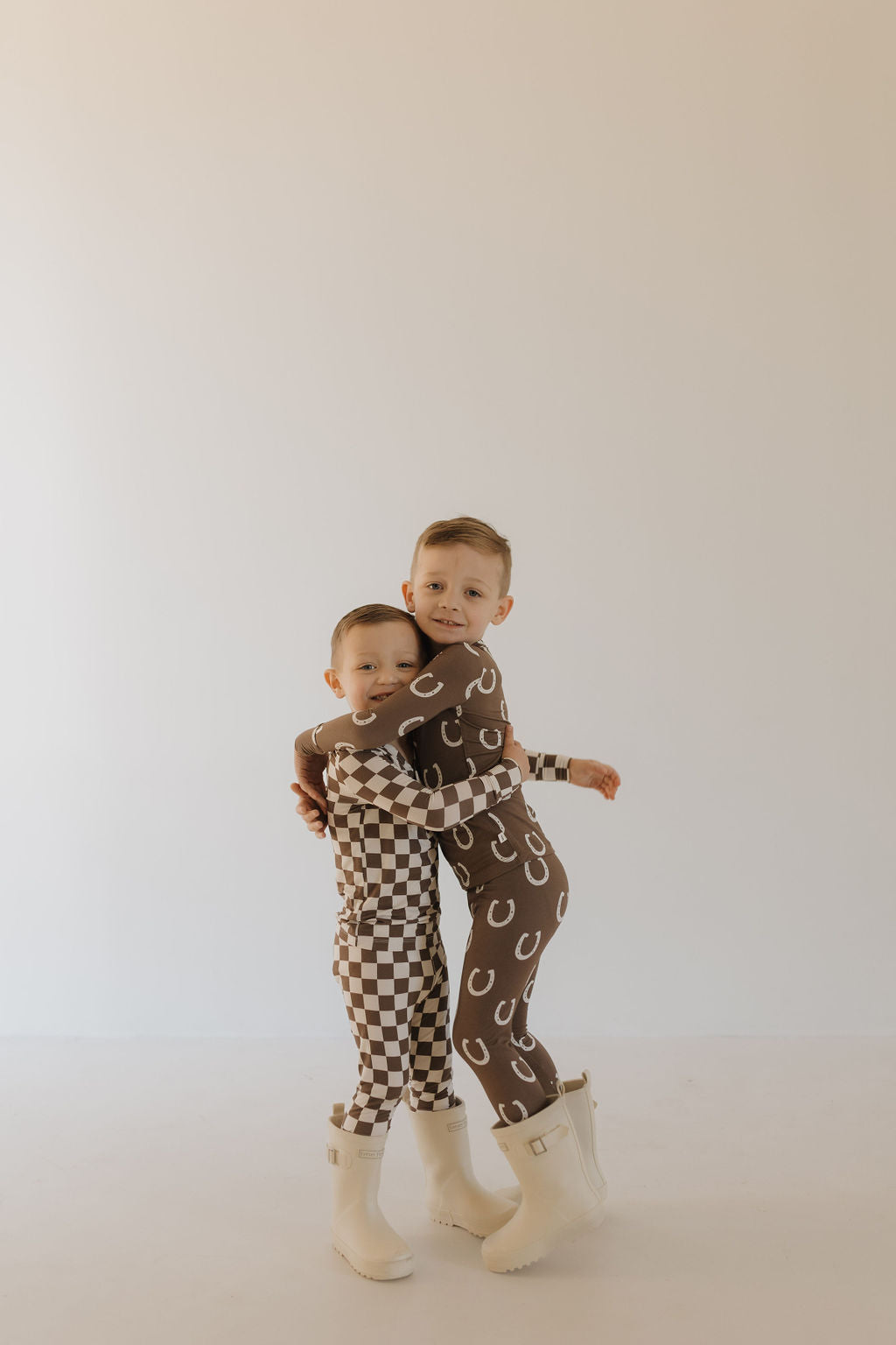 Two young children are joyfully hugging in their forever french baby outfits: one in Bamboo Two Piece Pajamas - Dutton with a white and brown checkered pattern, the other in brown with white crescents. Hypo-allergenic fabric ensures comfort, while white boots complement the scene on a plain backdrop.