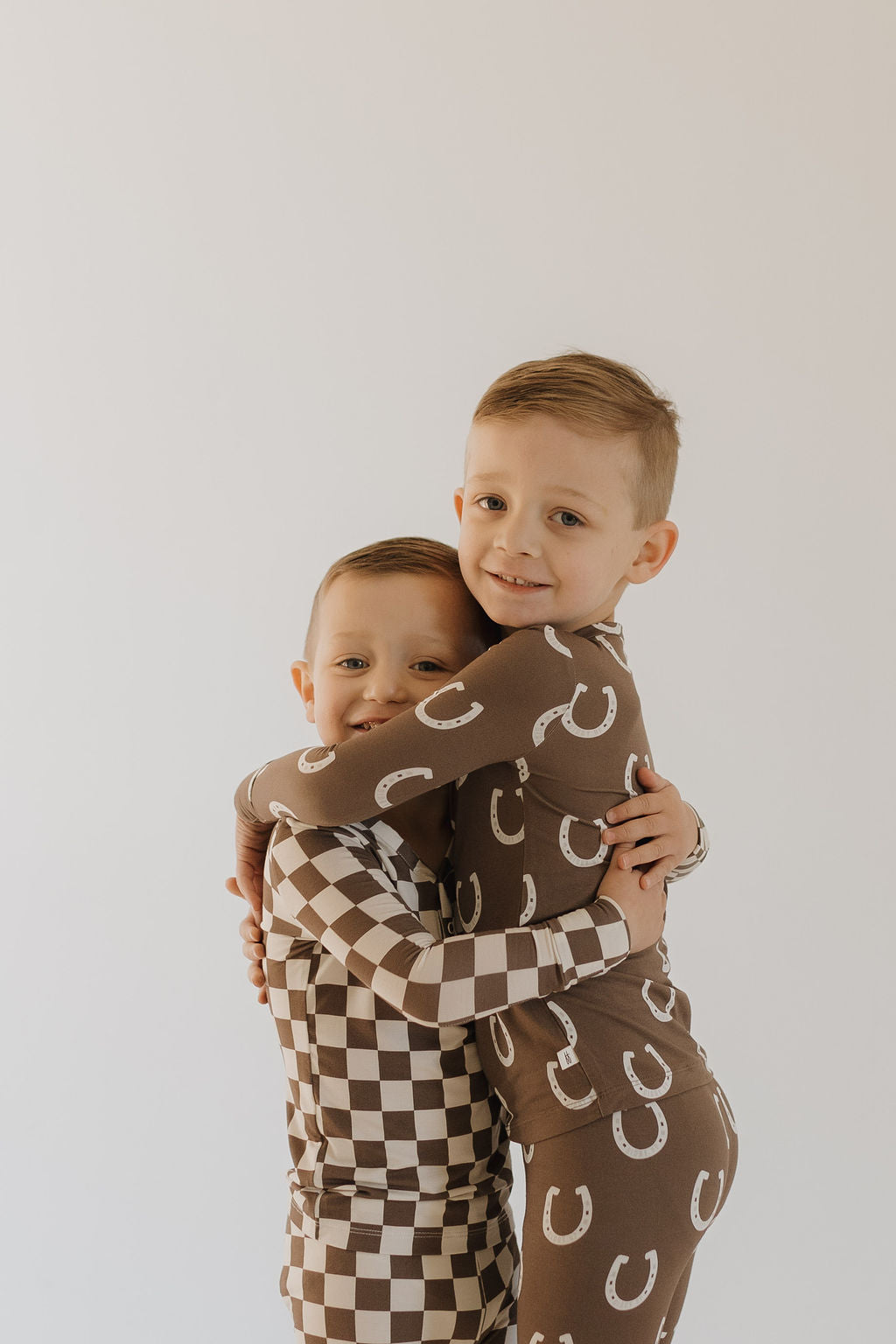 Two young children in forever french baby's Dutton bamboo two-piece pajamas share a hug. One wears a checkered pattern, and the other features the letter "C". These hypoallergenic, brown and white garments ensure comfort against a plain white backdrop.