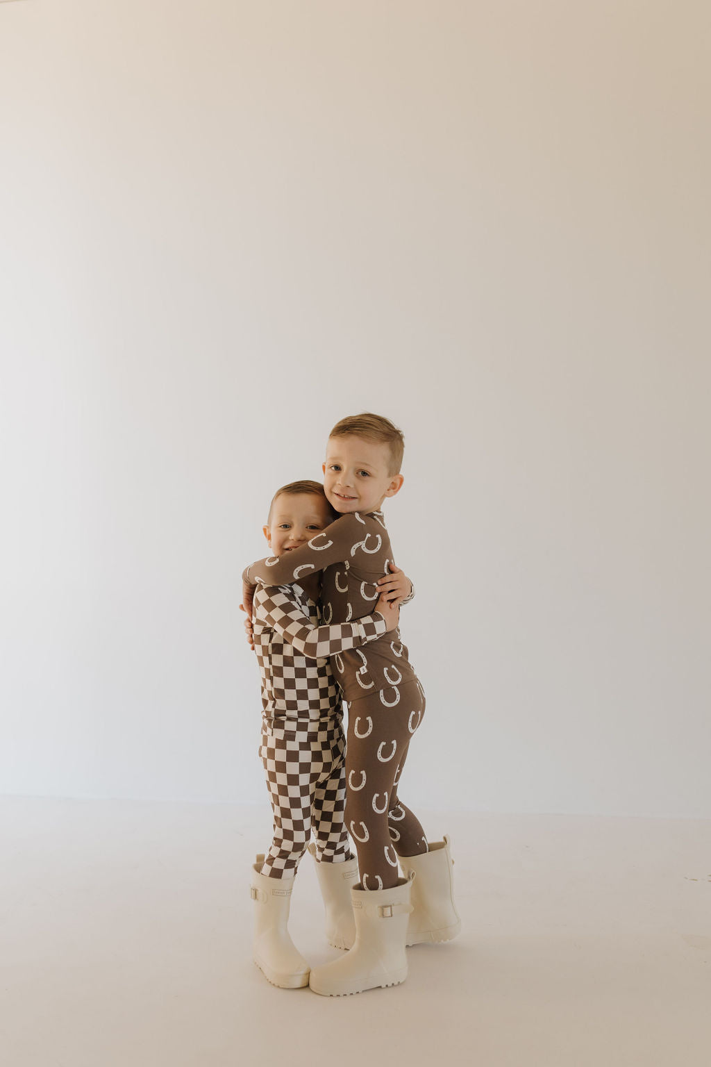 Two young children hug, wearing breathable forever french baby pajamas. One is in bamboo Dutton pajamas with a horseshoe pattern, while the other wears a brown and white checkered set. Both have white boots and stand against a light-colored backdrop.