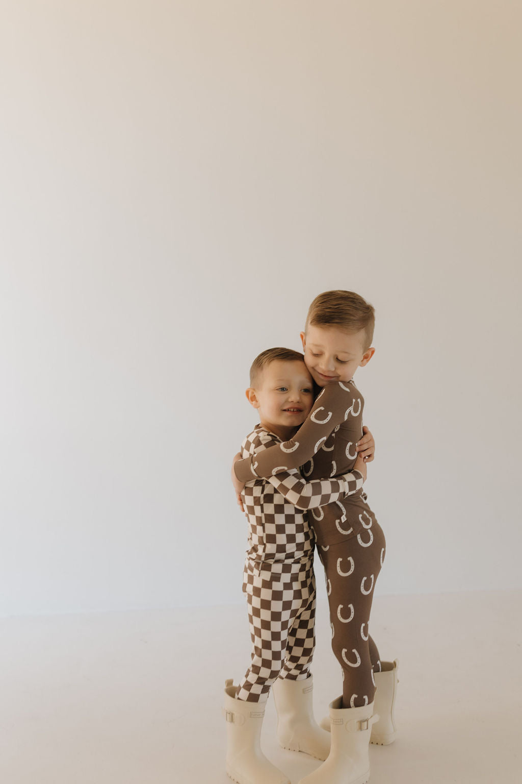 Two young children embrace in a neutral room. One wears breathable sleepwear with white horseshoe prints from forever french baby's Bamboo Two Piece Pajamas | Dutton, while the other sports a brown and white checkered outfit. Both smile happily, wearing white boots.