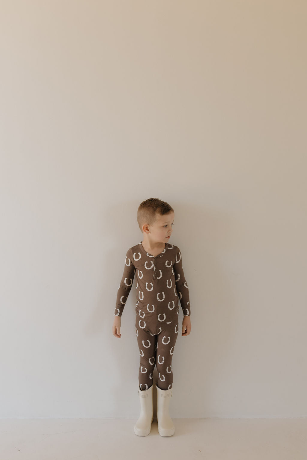 A young child in forever french baby's Bamboo Two Piece Pajamas, "Giddy Up," featuring a brown design with white horseshoe patterns, stands against a plain light-colored wall, looking to the side. The pajamas are crafted from breathable fabric and paired with white boots.