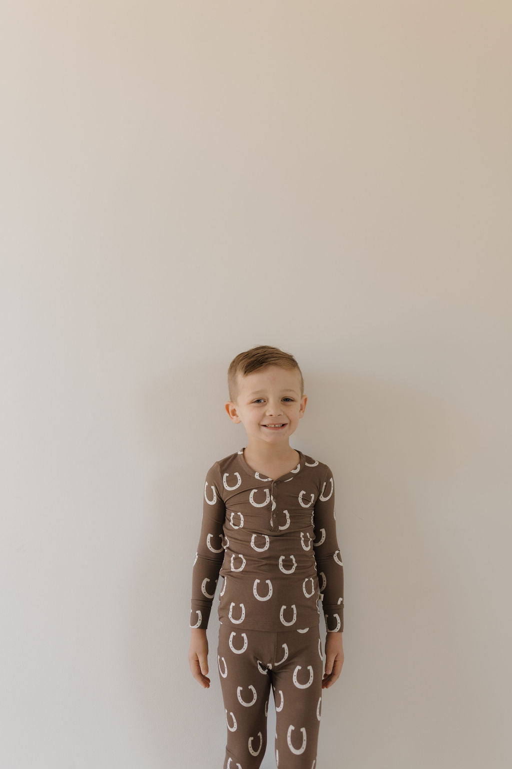 A smiling young child with short hair stands against a plain wall, wearing the forever french baby Bamboo Two Piece Pajamas in the Giddy Up pattern. These brown, hypoallergenic pajamas feature white horseshoes and are made from breathable fabric for ultimate comfort. The child looks directly at the camera.