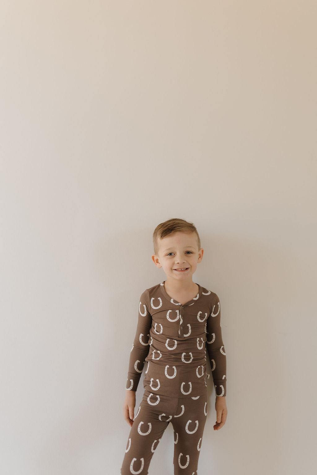 A young boy smiles against a light wall, wearing Bamboo Two Piece Pajamas in the Giddy Up style by forever french baby, featuring white horseshoe patterns. He enjoys the comfort of the breathable fabric.