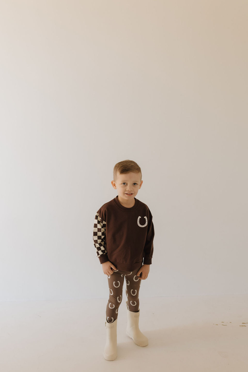 A child in the Forever French Baby Bamboo Two Piece Pajamas, Giddy Up design—featuring a brown horseshoe pattern and a checkered sleeve—stands against a plain background, paired with comfy beige boots.