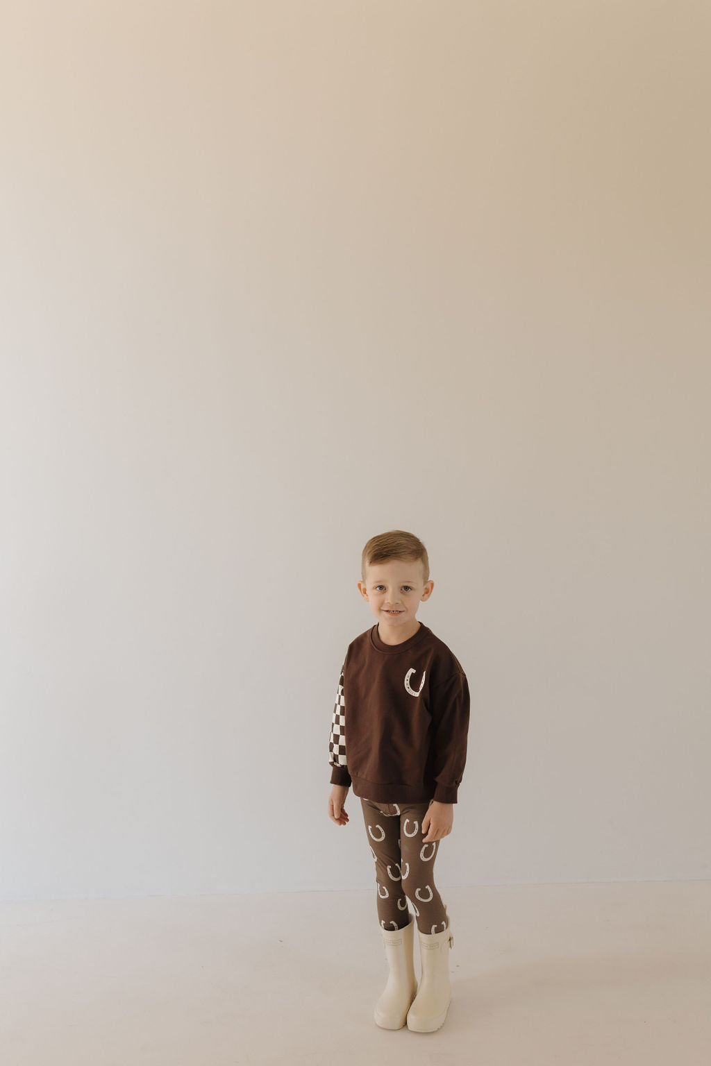 In a neutral-colored room, a young child wears the "Bamboo Two Piece Pajamas | Giddy Up" by Forever French Baby. The cozy attire features checkerboard sleeves and patterned pants in breathable fabric, paired with light boots and a neutral expression against the plain backdrop.