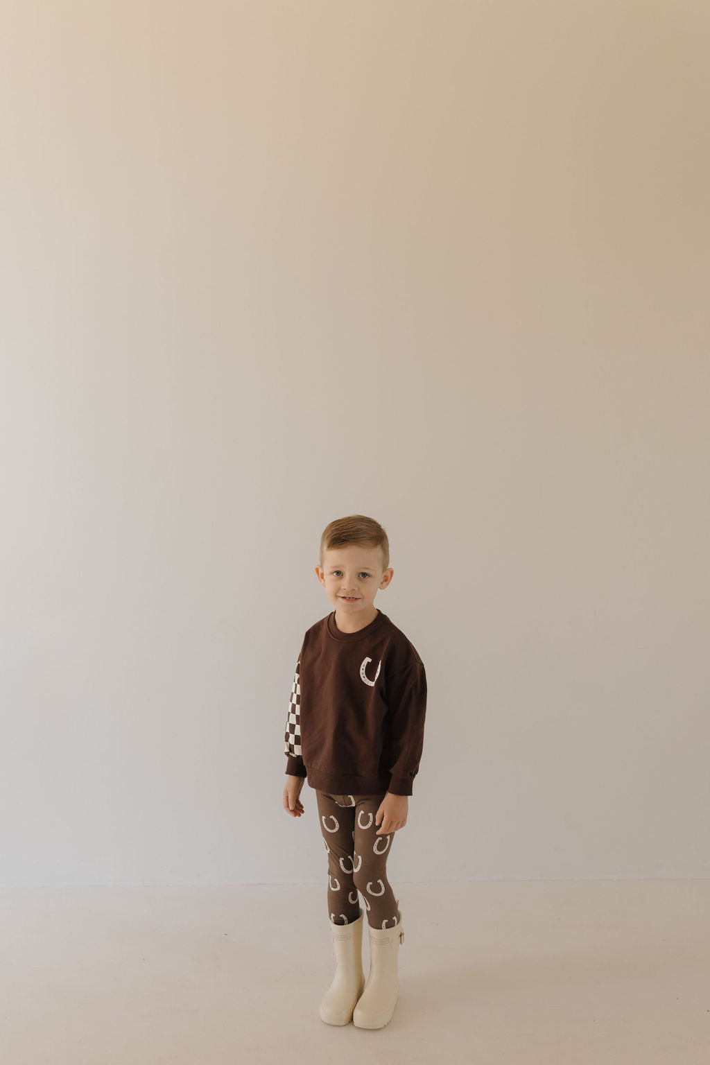 A young child stands indoors in a minimalistic setting, wearing forever french baby's breathable Bamboo Two Piece Pajamas in "Giddy Up," featuring a brown long-sleeve shirt with a crescent moon design and brown leggings with a horseshoe pattern, complemented by cozy white boots.