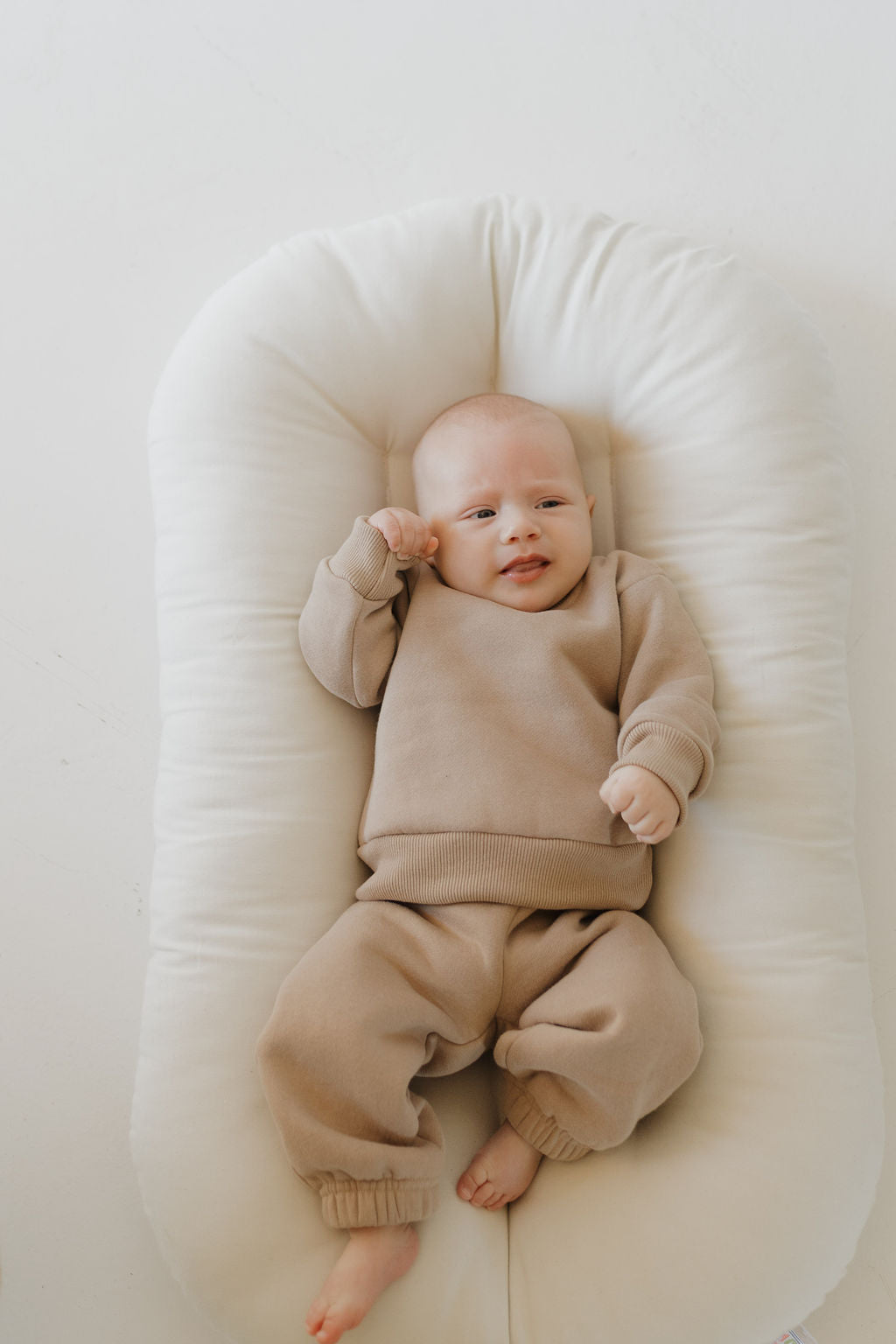 A baby wearing the forever french baby Sweat Set in Fawn relaxes on a cushioned white lounger. The soft material and elastic waistband allow easy movement as the baby looks up with one arm raised, enjoying the cozy surroundings.