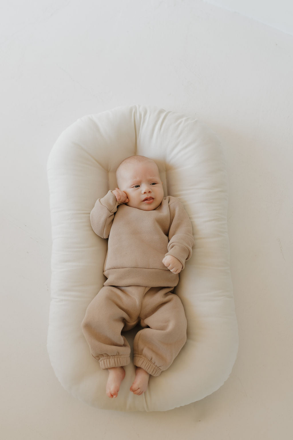 A baby wearing the "Sweat Set | Fawn" by forever french baby lies on a cushioned white pad. The super soft material ensures they appear relaxed with one hand near their face, set against a plain, white background.