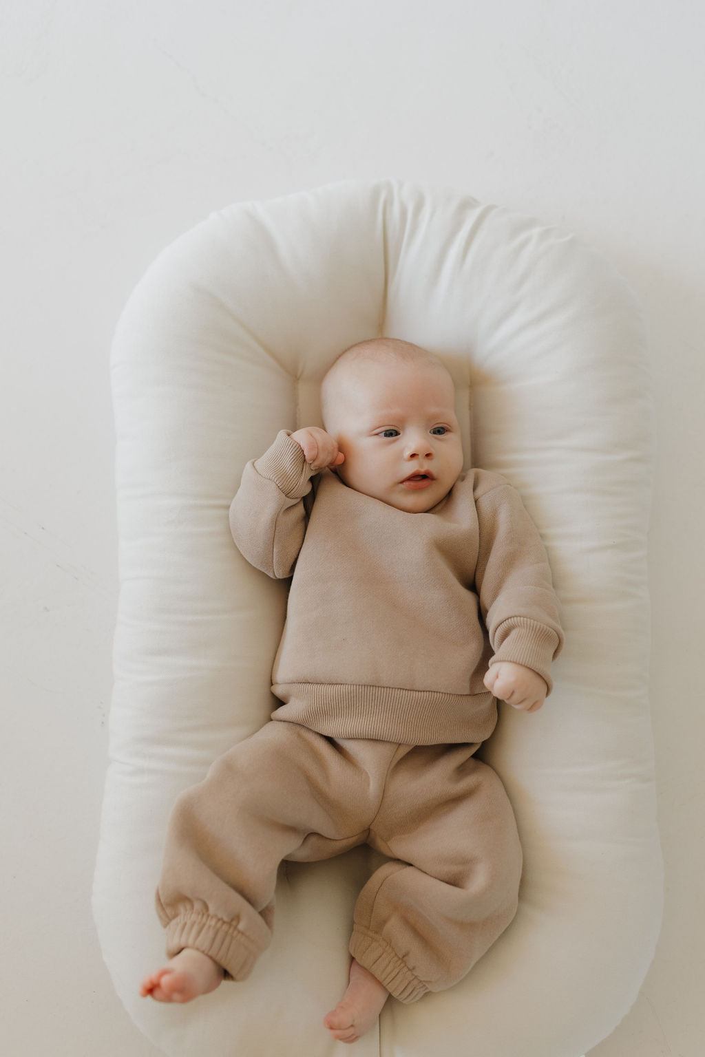 A baby wearing the "Sweat Set | Fawn" by forever french baby, with its elastic waistband and super soft material, rests contentedly on a plush white cushion, one arm raised against a neutral backdrop.