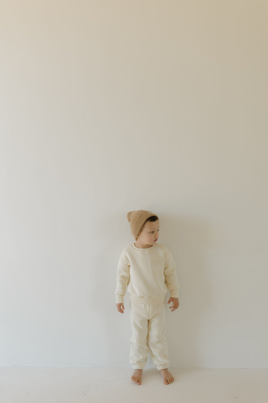 A child in a forever french baby "Sweat Set | Oat" stands barefoot on a light background, wearing an elastic waist outfit with a beige beanie. The minimalist setting uses soft neutral tones, and the material is super soft. They are gazing to the side.