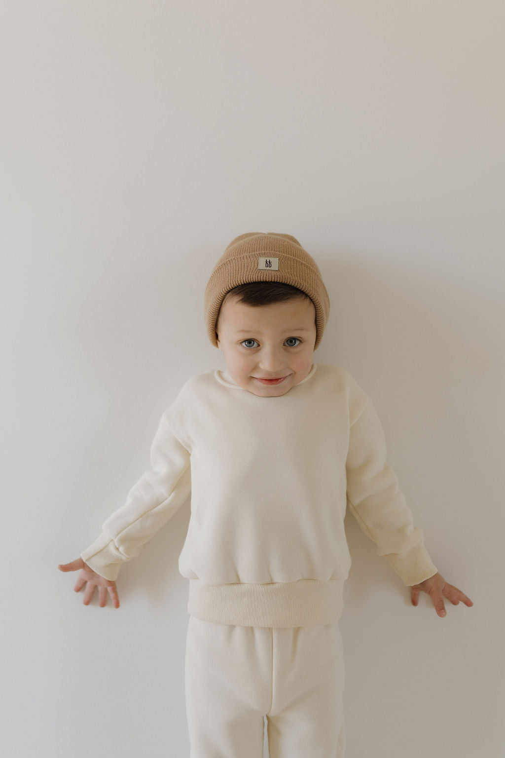 A child in a forever french baby Sweat Set | Oat, featuring an elastic waistband and matching knit beanie, stands against a white background with a playful expression and arms slightly outstretched to highlight the super soft material.