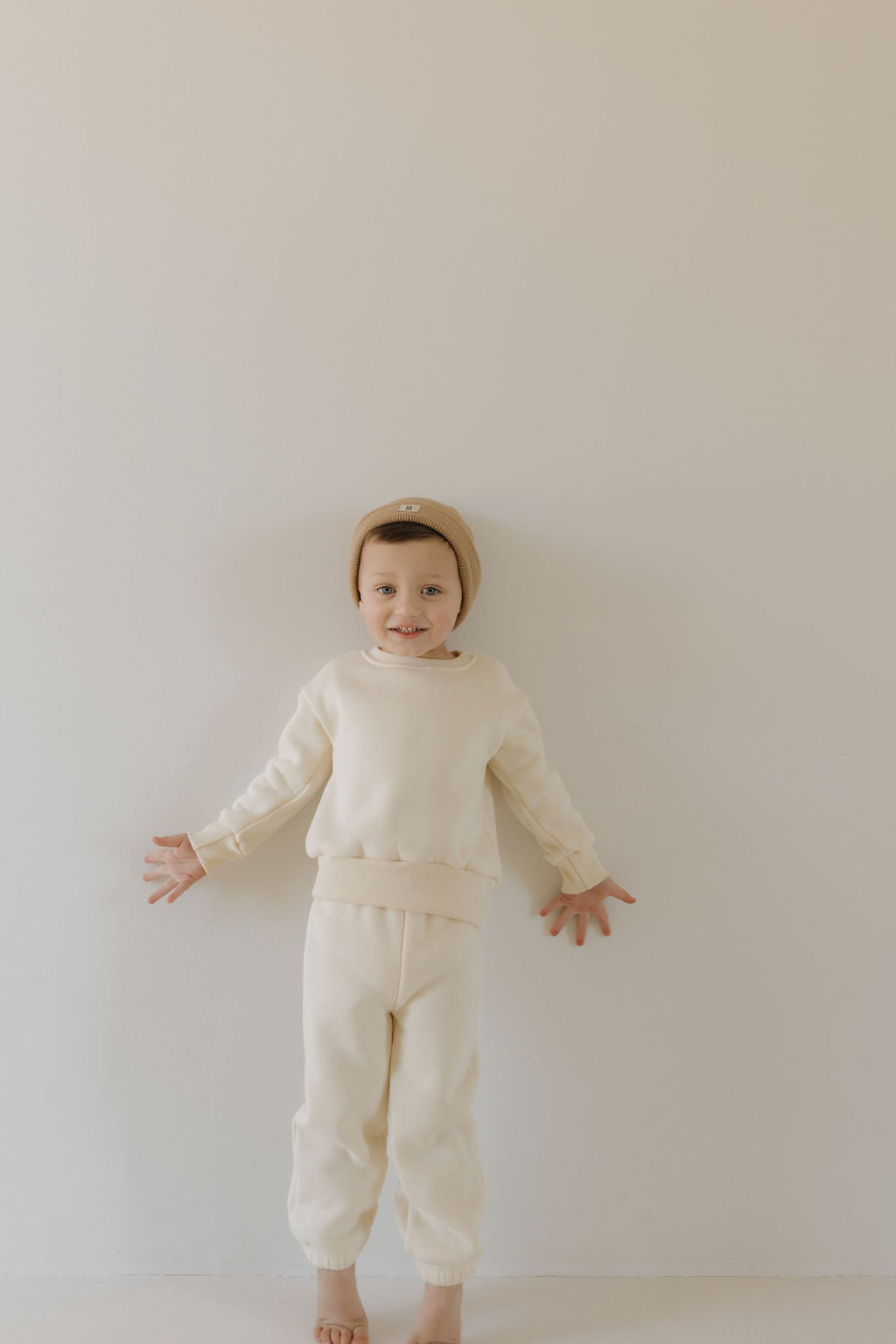 A cheerful child stands barefoot against a light background, wearing a cozy Sweat Set in Oat by forever french baby. The outfit features a super-soft material with a matching beanie and their arms slightly spread apart.