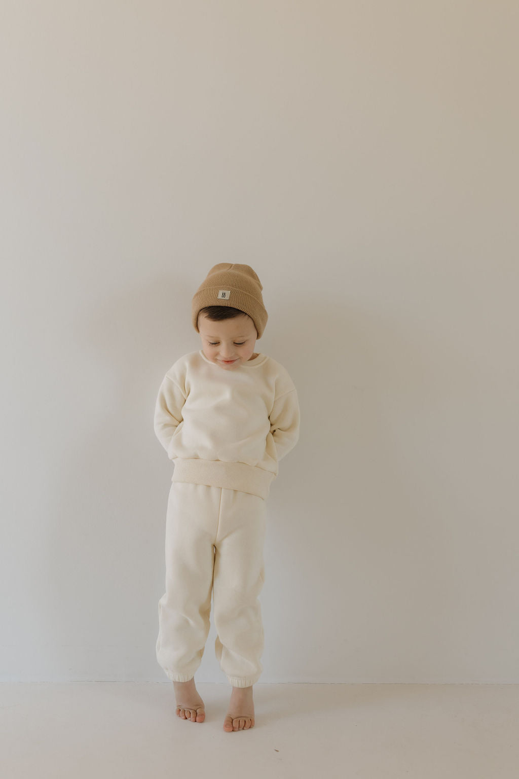 A child stands barefoot against a light wall, wearing the forever french baby Sweat Set | Oat. This cozy light beige sweat set, featuring a super soft sweatshirt and matching pants, pairs perfectly with a brown beanie as the child gazes down with hands behind their back.