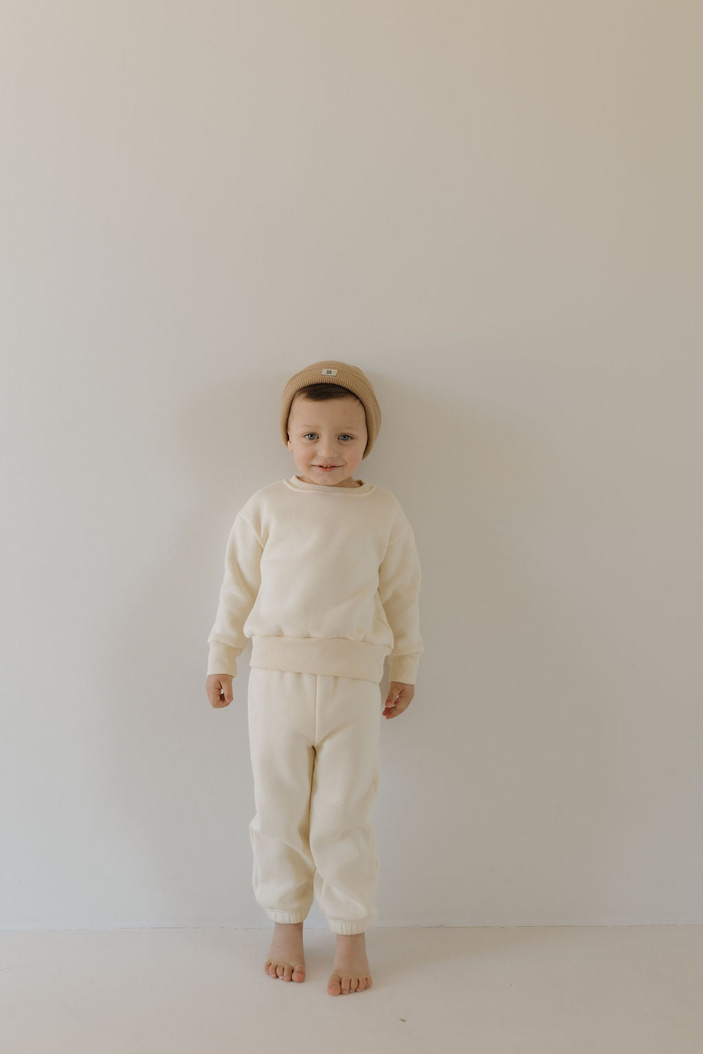 A small child poses against a plain light background, wearing the forever french baby Sweat Set in Oat with an elastic waistband. A brown knit beanie complements their outfit, and they are without shoes. The super soft material enhances their calm and neutral expression.