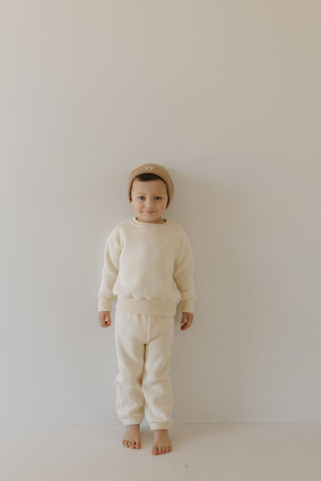 A child poses against an off-white backdrop wearing the "Sweat Set | Oat" by forever french baby. Complemented by a beige beanie and bare feet, the outfit's elastic waistband enhances their relaxed vibe as they gently smile at the camera.