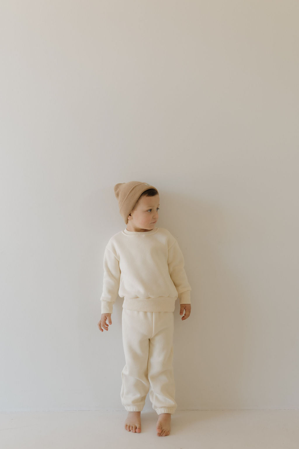 A young child is against a plain wall in the "Sweat Set | Oat" from forever french baby, featuring a cozy light beige sweater and matching pants with an elastic waistband, paired with a brown beanie. They look to the side, their bare feet touching the light-colored floor.