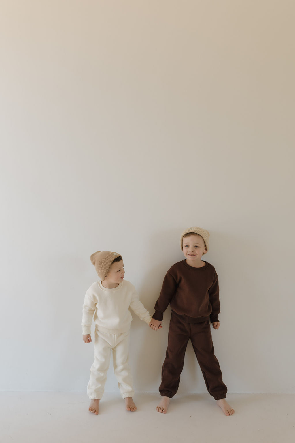 Two young children hold hands against a neutral background. One wears a cream outfit and beanie, while the other sports the forever french baby Sweat Set in Driftwood with an elastic waistband. Both are barefoot, content, and enjoying their all-day comfort together.