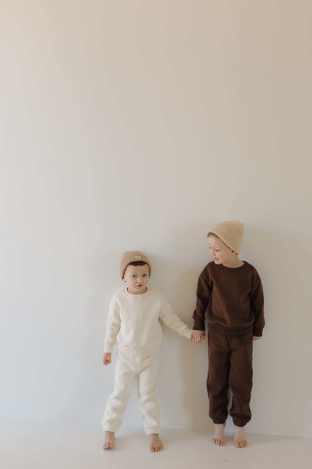 Two young children stand barefoot against a plain background, holding hands. One wears a white outfit with an elastic waistband, the other in the "Sweat Set | Oat" by forever french baby made from super soft material. Both wear knitted beanies and have serene expressions.