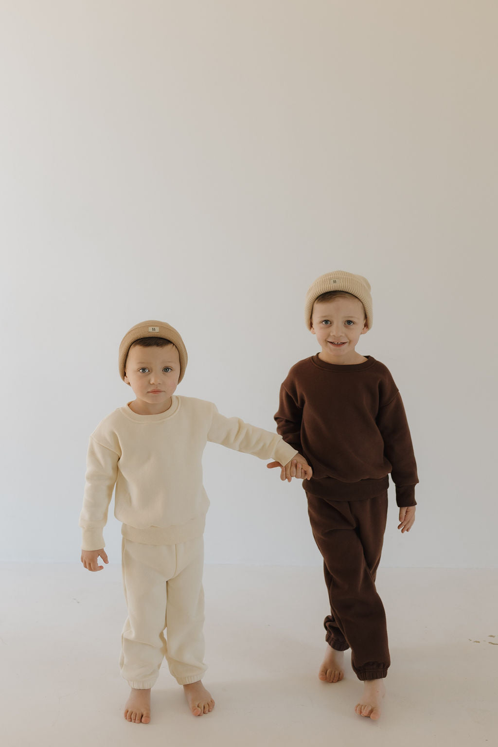 Two young children in beanies stand barefoot on a plain floor against a neutral background. One wears forever french baby's Sweat Set | Oat, while the other sports a brown outfit made of super soft material. Holding hands, the child in brown stands slightly ahead.