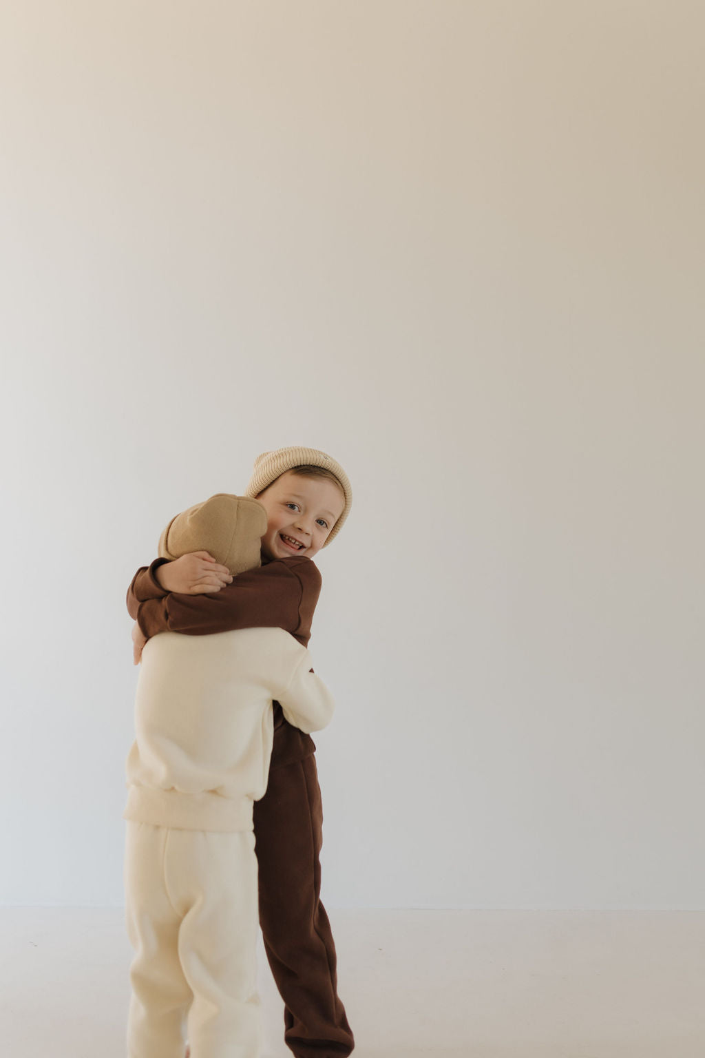 Two children hug warmly in a minimalist studio. One wears a brown outfit and beanie, while the other is dressed in forever french baby's "Sweat Set | Oat," made from super soft material. The light-toned background complements their cozy attire.