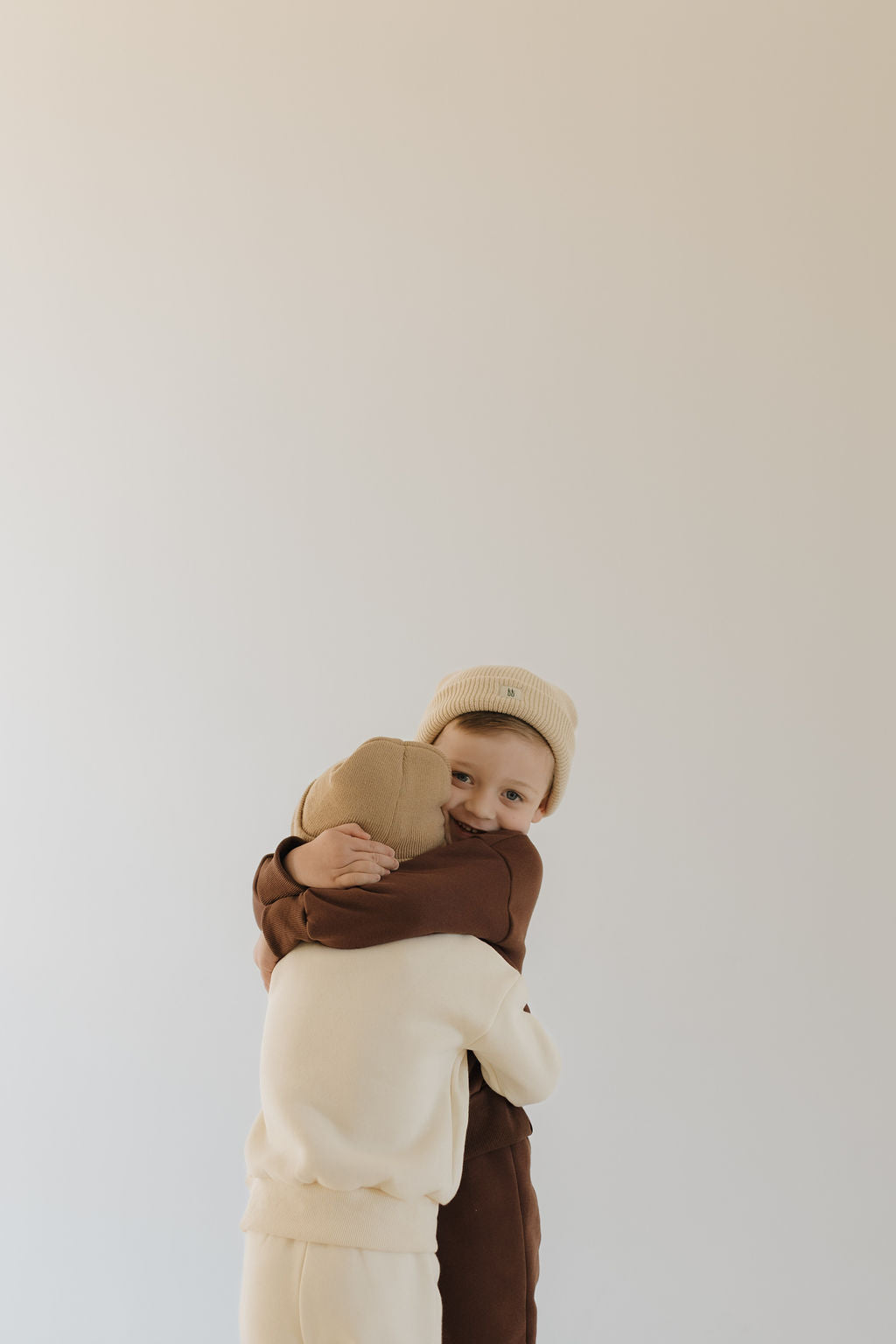 Two children are hugging, enveloped in the all-day comfort of forever french baby's Driftwood sweat set. One wears brown with a beige hat, the other cream with a tan hat, while a plain background highlights their warmth and love.