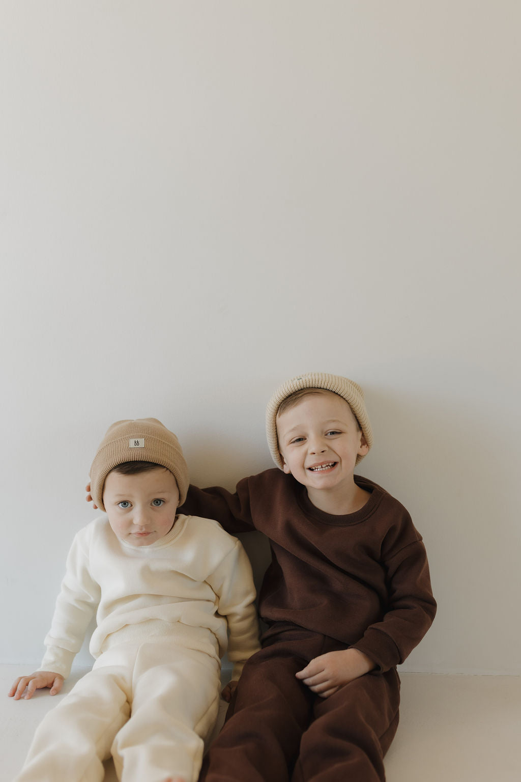 Two young children sit against a plain white wall, both wearing matching beige beanies. The child on the left is in a cream outfit, while the one on the right wears a cozy "Sweat Set | Oat" from forever french baby. With an arm gently around their friend, the latter beams with joy.