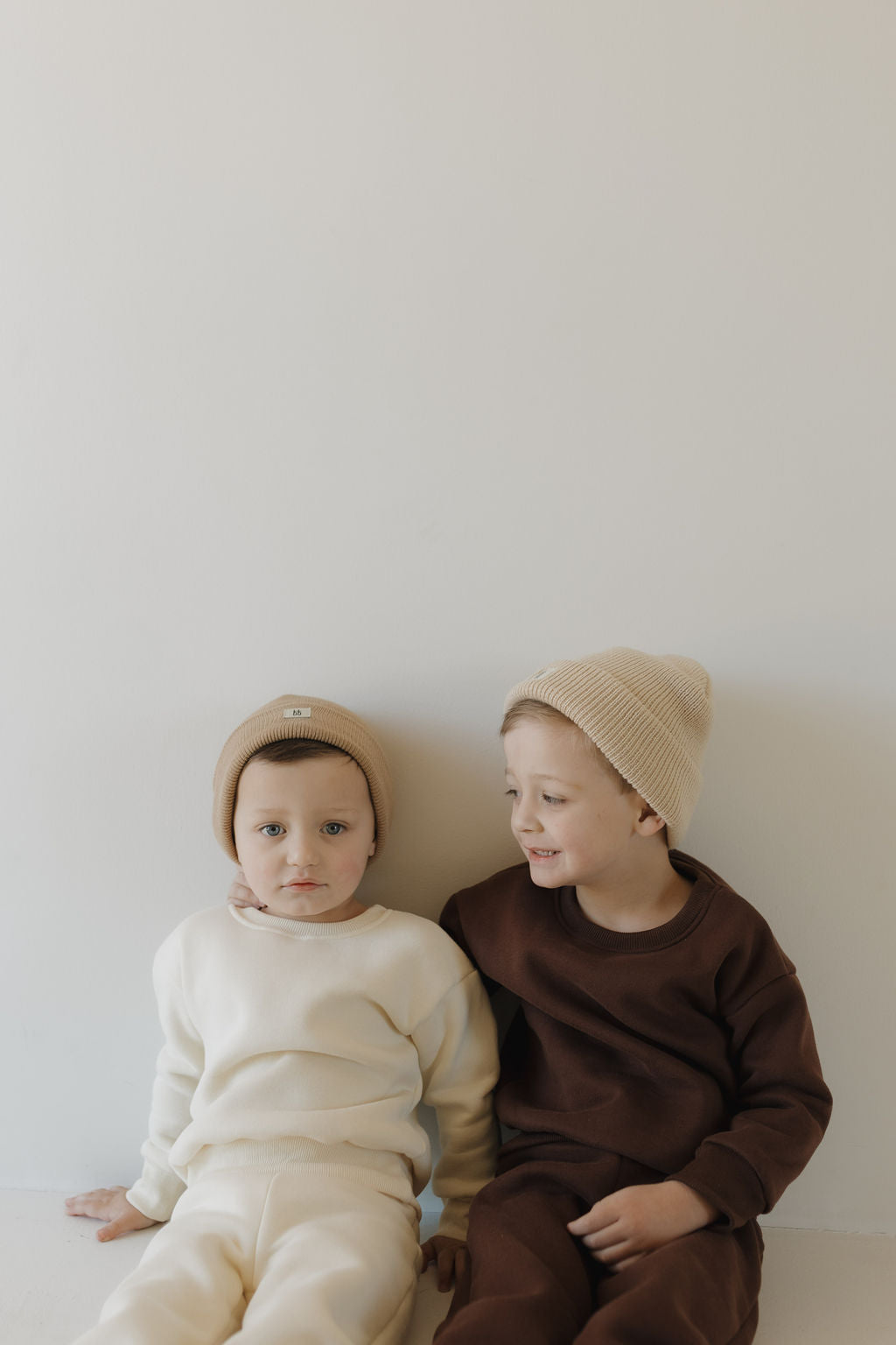 Two young children sit against a plain wall in cozy sweat sets by forever french baby. The child on the left wears cream with a beanie, while the other smiles warmly in a Driftwood brown ensemble featuring an elastic waistband for all-day comfort, seated close together in contentment.