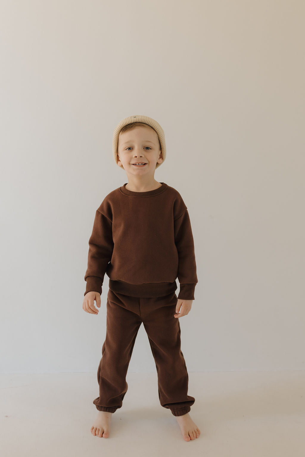 A young child is barefoot against a plain background, smiling in a Driftwood sweat set by forever french baby, featuring an elastic waistband for comfort. They complete the look with a beige knit beanie.