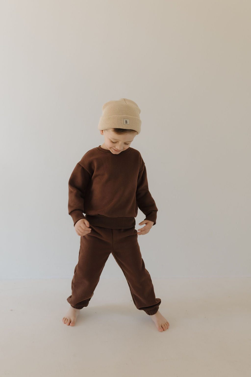 A young child in a cozy Driftwood Sweat Set by forever french baby stands barefoot on a light floor, wearing a beige beanie with a playful expression. The plain, light background enhances the minimalist setting, ideal for all-day comfort.