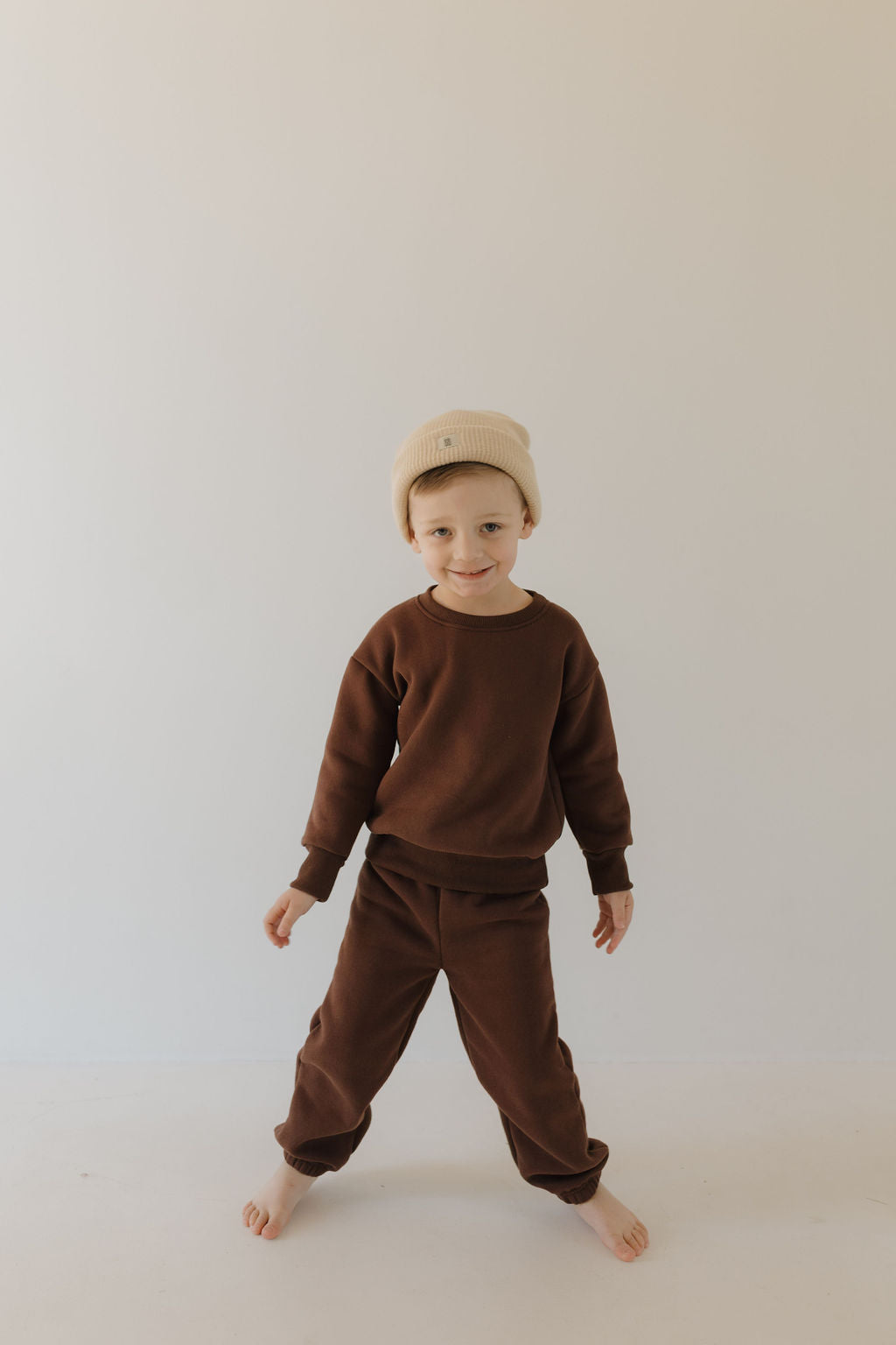 A young child poses barefoot against a plain backdrop, wearing the Driftwood Sweat Set by forever french baby. The brown sweater and elastic waistband pants are paired with a beige beanie, creating a cozy look as they smile brightly with arms relaxed, embodying all-day comfort.