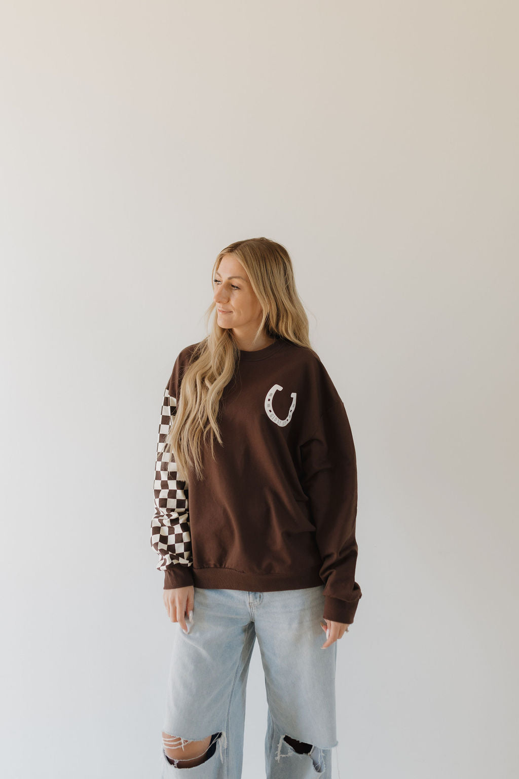 A person with long blonde hair stands against a light background wearing the "Giddy Up" brown sweatshirt from the forever french baby Howdy Collection, featuring a horseshoe design and checkered sleeves, paired with ripped jeans. This cozy piece creates a perfect matching moment as they gaze aside.