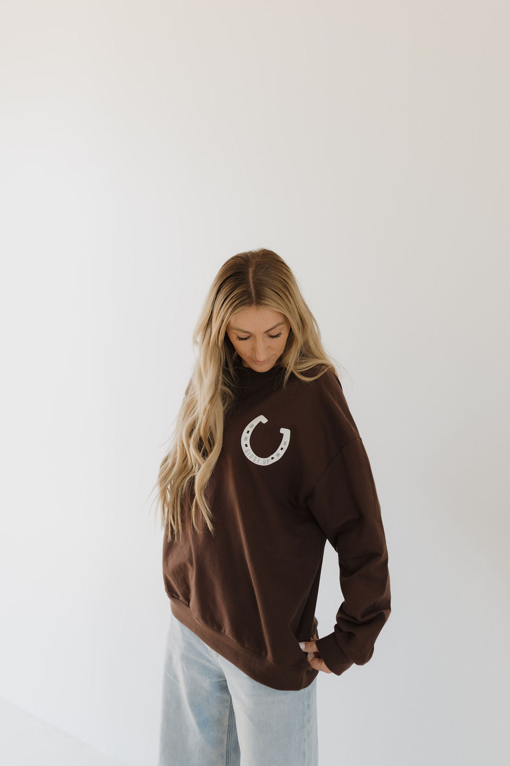 A person with long hair sports the forever french baby Adult Sweatshirt in brown from the Giddy Up line, showcasing a horseshoe design. They pair it with light-colored pants for a coordinated look while gazing down against a plain white backdrop.