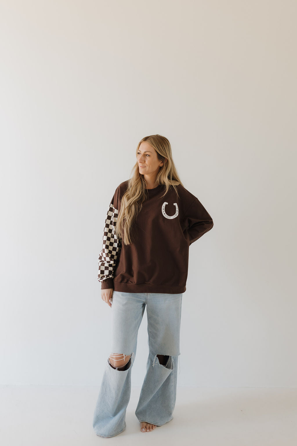 Against a plain background, a person with long hair embodies a stylish moment in the Adult Sweatshirt | Giddy Up from forever french baby. The brown top, adorned with a horseshoe emblem and checkered sleeves, pairs perfectly with light blue ripped jeans as they confidently place one hand on their hip.